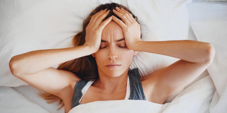 Niedobór jakiej witaminy powoduje bezsenność? Kobieta z zamkniętymi oczami leży w łóżku i trzyma dłonie na swoich skroniach.