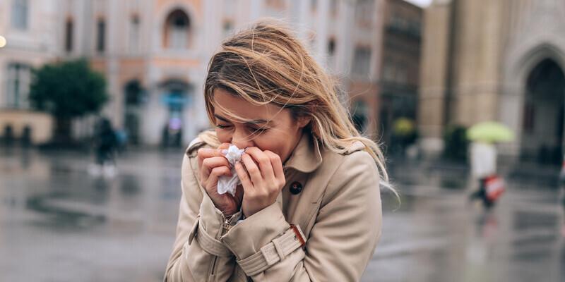 Jak szybko wyleczyć przeziębienie? Kobieta idąca przez miasto kicha, trzymając chusteczkę przy twarzy.