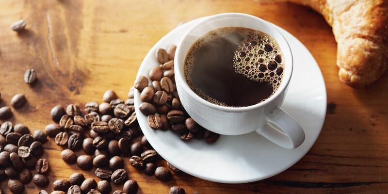 nadwrażliwość na kofeinę - zdjęcie przedstawia zaparzoną kawę w filiżance, kawę w ziarnach rozsypaną na stole oraz rogalika